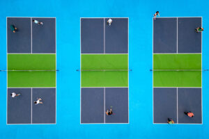 Aerial,View,Of,Multiple,Pickleball,Courts,With,Players,In,Action. Aerial view of multiple pickleball courts with players in action. Colorful blue and green outdoor sports complex on a sunny day. Recreational activity and healthy lifestyle concept